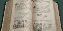 Das Logo zeigt ein aufgeschlagenes historisches Buch mit dem Text "Sacra Scripta" darunter.